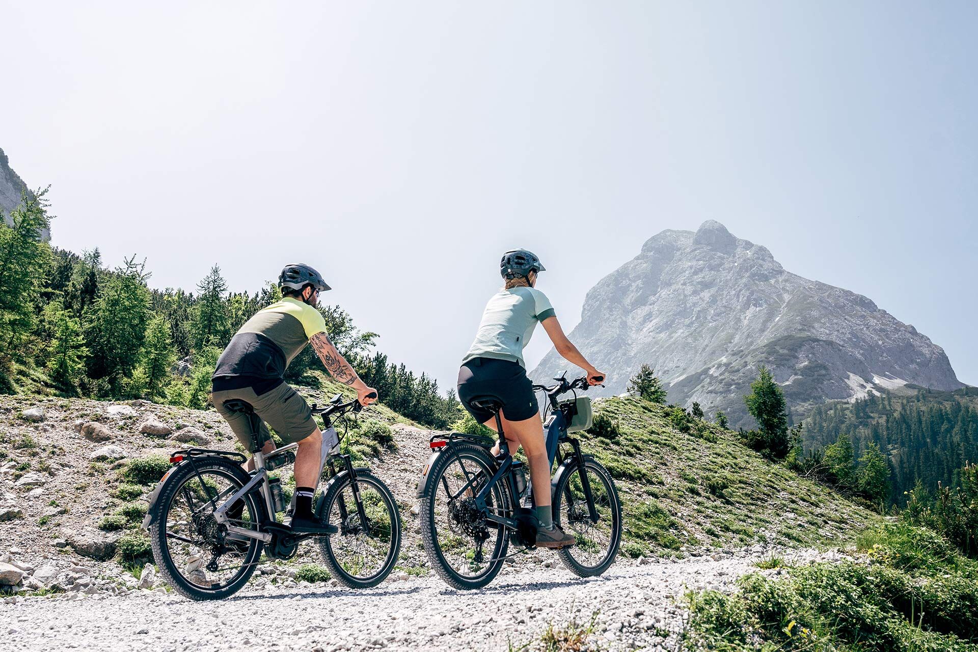 Un couple roule en montée sur un chemin de gravier avec des vélos électriques SUV. On aperçoit un sommet de montagne en arrière-plan.