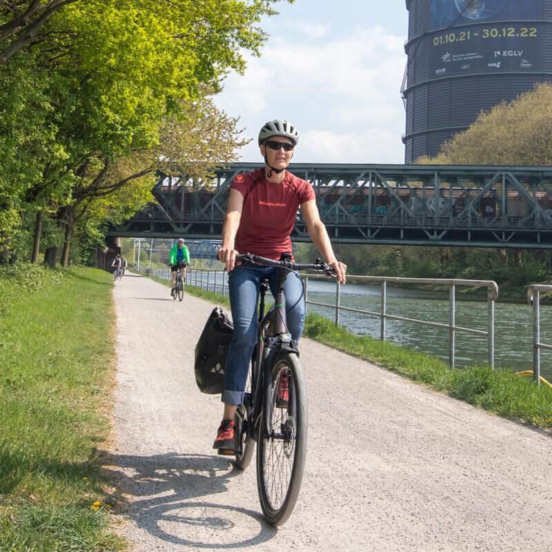 Frau fährt mit dem E-Bike den Rhein-Herne-Kanal entlang, im Hintergrund der Gasometer