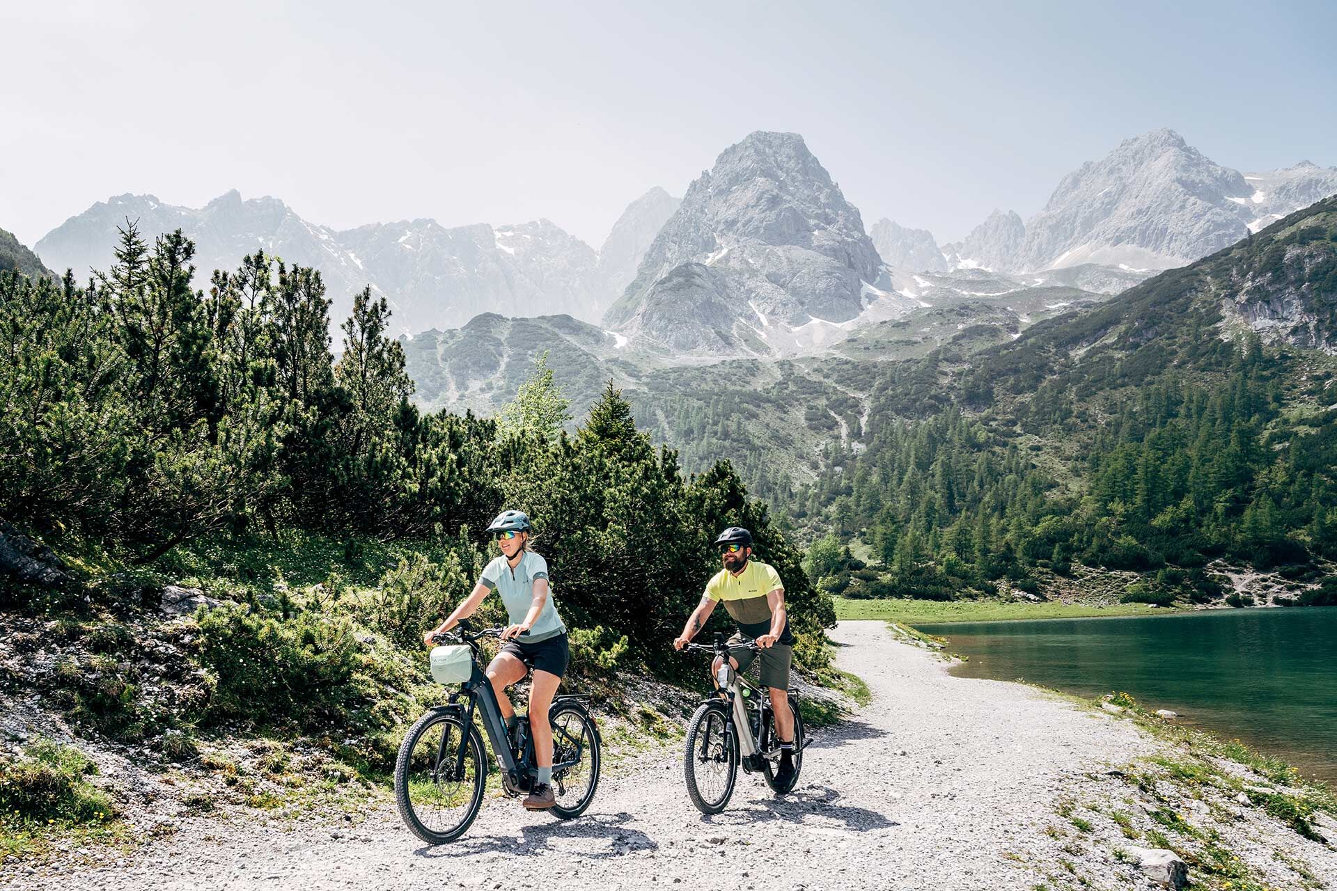 Un couple roule sur un chemin longeant les rives d'un lac de montagne, avec un panorama montagneux en arrière-plan.