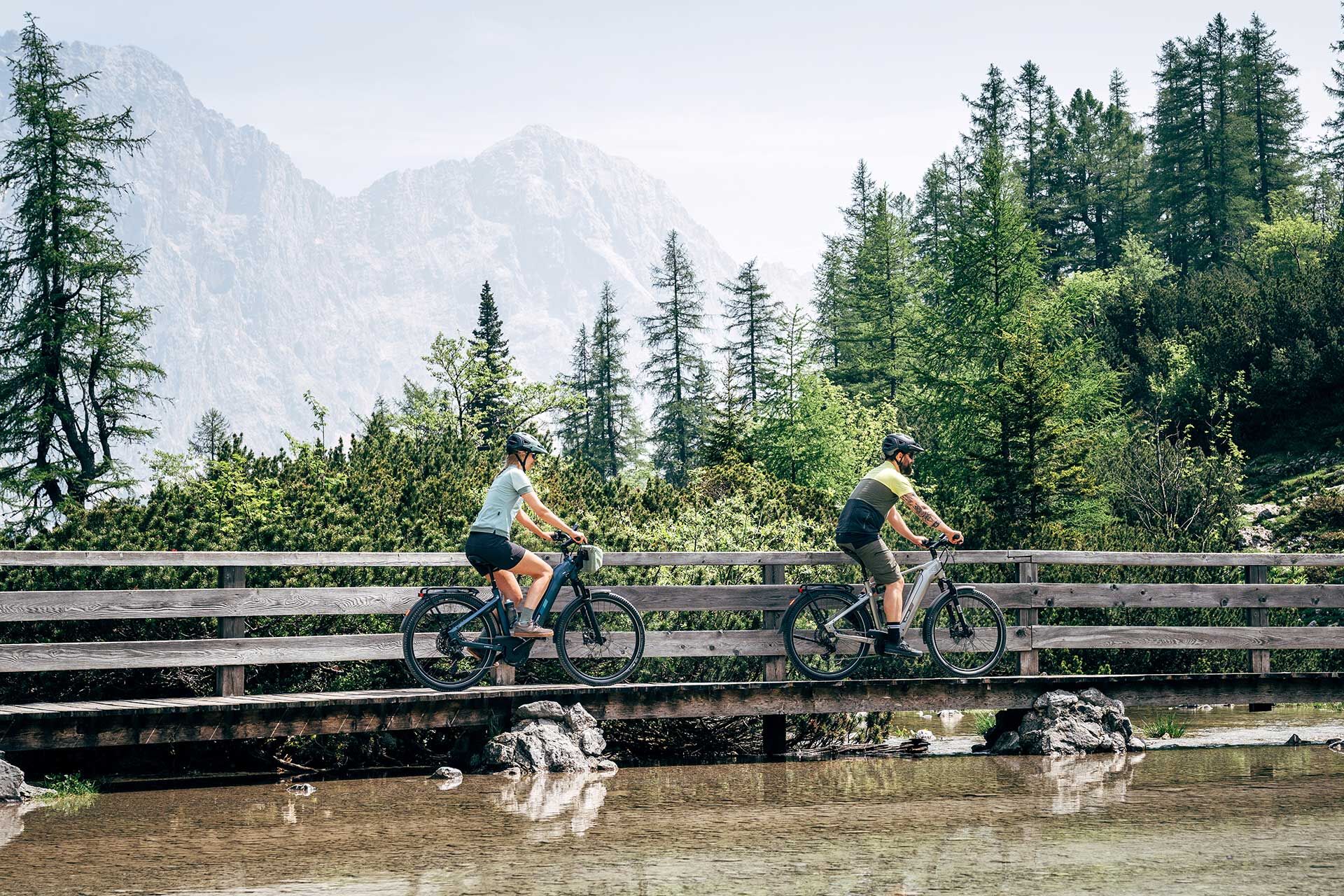 Un couple traverse un pont sur une rivière à vélo électrique, avec des montagnes en arrière-plan.