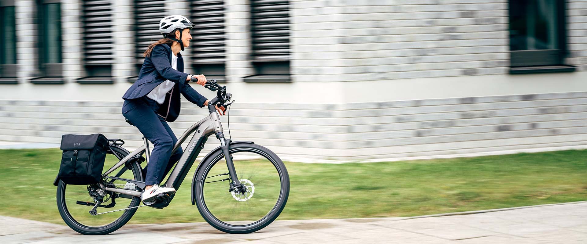 Eine Frau in Anzug und Helm fährt mit einem Elektrofahrrad, an dem eine schwarze Tasche befestigt ist, an einem modernen Gebäude vorbei.