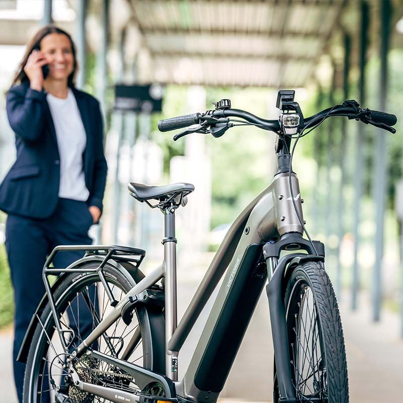 Ein Trekking E-Bike in einer urbanen Umgebung, im Hintergrund steht eine Frau im Anzug und telefoniert