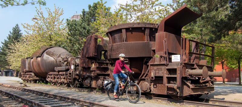 Eine Frau fährt mit dem E-Bike an einem alten, stillgelegten Zug vorbei