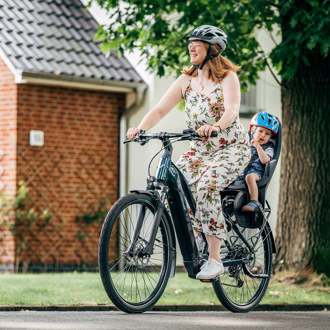 Eine Frau fährt mit einem E-Bike durch eine Vorstadtsiedlung. Am Gepäckträger sitzt ein Kind im Kindersitz.